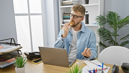 Handsome young man with beard eating in cluttered office, surrounded by technology and plants.
