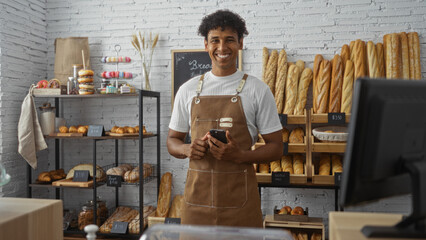 Fototapeta premium Handsome young man in apron standing in bakery shop surrounded by fresh bread smiling at camera