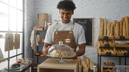 Young man in an apron smiling while holding a cake stand filled with pastries in a bakery shop interior