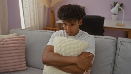 Obraz premium Young hispanic man sitting in a living room hugging a pillow while looking pensive in a modern apartment interior