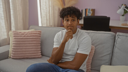 Fototapeta premium Young man sitting on a couch in a cozy living room, contemplating deeply with a thoughtful expression in a modern apartment setting.