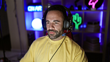 Portrait of a focused hispanic man with a beard wearing headphones in a colorful gaming room during night time.