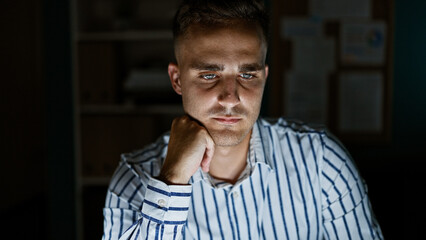 Obraz premium Thoughtful young hispanic man in a striped shirt posed in a dimly lit office room, evoking a sense of contemplation.