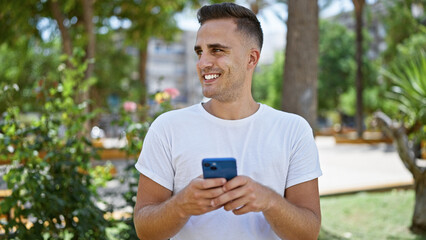 Fototapeta premium A cheerful young hispanic man texting on his smartphone while enjoying the sunny ambiance in a lush city park.