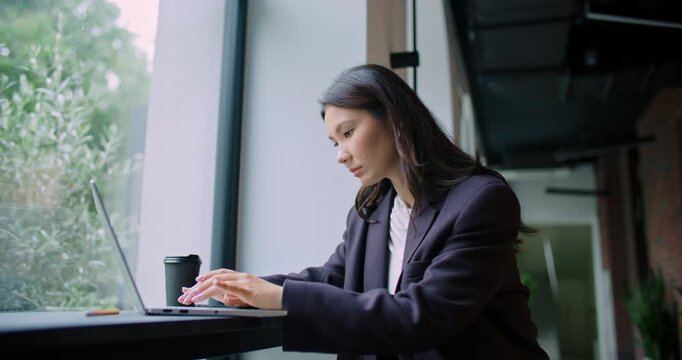Charming Asian woman sitting at thin desk in front of large window. Typing very fast on keyboard. Entering information or searching for data online. Cup of hot drink next to computer.