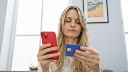 Fototapeta premium Concerned young woman holding credit card and smartphone in modern living room.