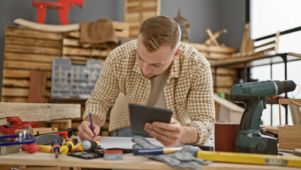 Bearded man using tablet and planning in a woodwork studio surrounded by tools and lumber.