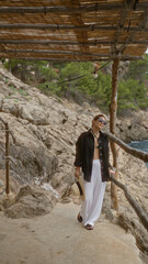 Woman relaxing outdoors at cala deia in mallorca, spain, wearing sunglasses and holding a hat, surrounded by rocky terrain and greenery under a rustic wooden shelter.