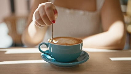 Cafe bliss, young woman enjoys leisurely coffee break, reveling in the sunny interior of a cafeteria, sitting, delicately stirring her drink indoors, closeup showcasing the relaxation