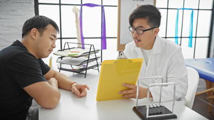 A male doctor in a white coat discusses with a male patient in an office with a spine model in the...