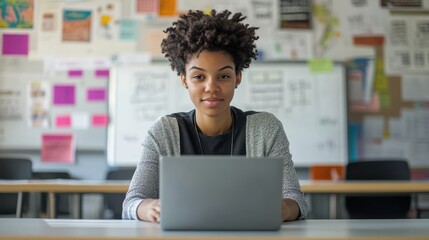 A young teacher sitting in a modern classroom, working on a laptop, with a whiteboard and educational posters in the background, highlighting the blend of traditional and digital teaching methods