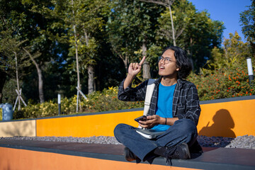 A man wearing a plaid shirt sitting in the park makes an expression of getting an idea.