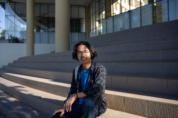 Fototapeta premium A male student wearing a plaid shirt sits on the stairs and smiles