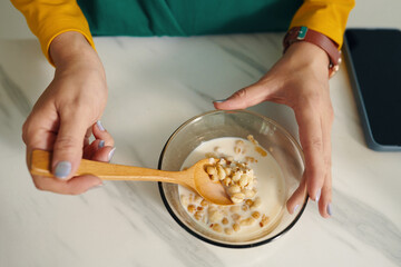 Hands adding nuts to healthy breakfast cereal using wooden spoon. This close-up image captures detailed process showcasing nutritious meal preparation on marble countertop