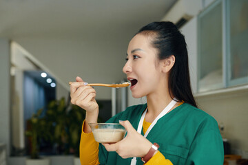 Asian health care professional enjoying a meal in clinic setting while wearing scrubs and holding a...