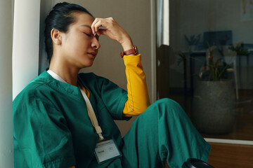 Tired Asian healthcare worker leaning against wall wearing green and yellow scrubs with closed eyes. ID badge visible on person, suggesting role as medical professional