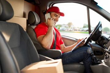 Delivery service. Young courier man talking on cellphone and looking at clipboard, checking address of customer, sitting in van