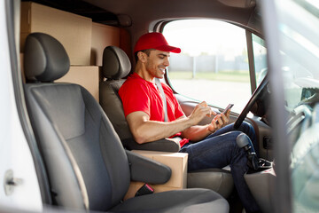 Young positive deliveryman courier sitting in van and using smartphone, texting to customer or...