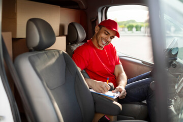 Young smiling courier man checking amount of parcels and writing information in clipboard, sitting in delivery van