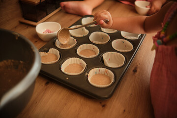 A close-up of a child's hands filling cupcake molds with batter, surrounded by baking ingredients in a modern kitchen, emphasizing creativity and fun family activities.