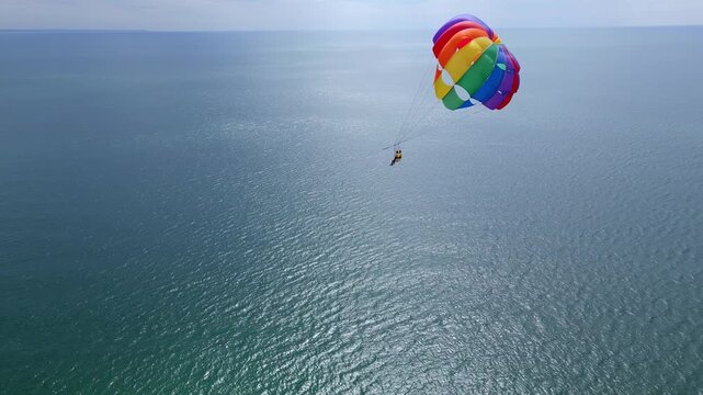 Drone video of a colorful parasail soaring over the open ocean, towed by a speedboat below. The aerial view captures the thrill and freedom of parasailing against a vast, serene seascape