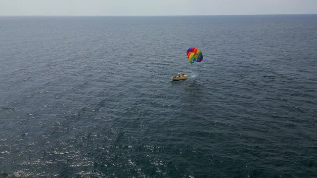 Drone video of a colorful parasail soaring over the open ocean, towed by a speedboat below. The aerial view captures the thrill and freedom of parasailing against a vast, serene seascape