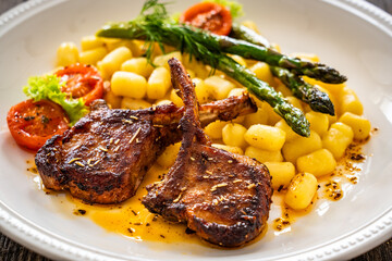 Lamb chops with bone, gnocchi and cooked green asparagus served on white plate on wooden table 