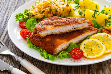 Crispy breaded fried pork chop, boiled potatoes and sauerkraut salad served on white plate on wooden table 