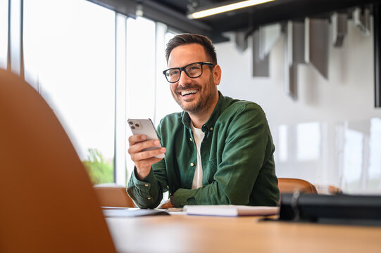 Cheerful handsome businessman in eyeglasses looking away while using mobile phone at desk in office