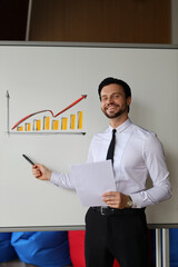 A man is standing in front of a white board with a graph on it. He is holding a piece of paper and pointing to the graph. The man is smiling and he is giving a presentation