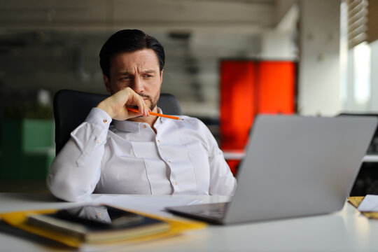 A man is sitting at a desk with a laptop and a pencil. He is looking at the laptop screen and he is deep in thought