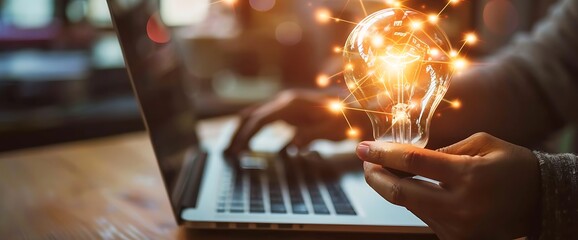 A hand holds a glowing light bulb with sparkling lines emanating from it, suggesting a new idea, innovation, or inspiration, in front of a laptop computer.