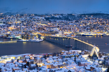 Obraz premium Panorama of Norwegian city of Troms in the winter. Snowy roofs, bridge, embankment near the port and fishing ships. Evening lights