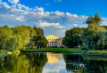 Fototapeta premium A breathtaking view of a grand manor house reflecting in a serene pond, surrounded by lush greenery and under a vibrant blue sky. The peaceful landscape captures the essence of tranquility and