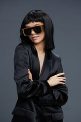 Businesswoman in a black suit with sunglasses and a gold chain poses confidently against a gray background