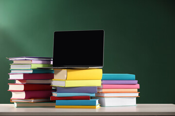Stacks of many colorful books and laptop on white wooden table near chalkboard in classroom