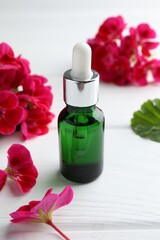 Bottle of geranium essential oil and beautiful flowers on white wooden table, closeup