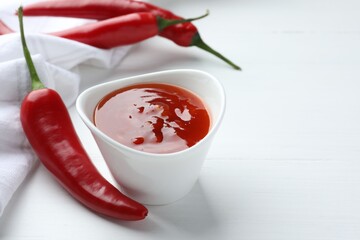 Spicy chili sauce in bowl and fresh peppers on white wooden table, space for text