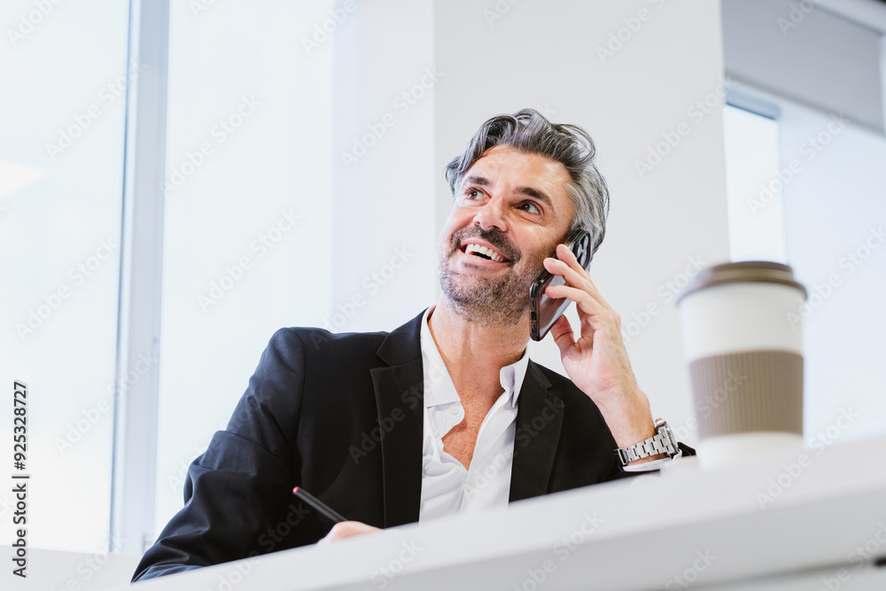 Wall mural From below of cheerful, grey-haired executive enjoys a pleasant conversation on the phone while writing notes in a sunlit, modern office