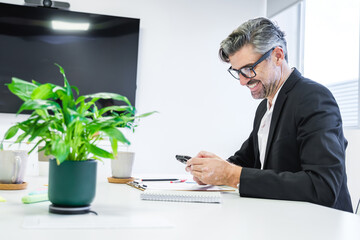 A genial executive in a sleek suit is seen engaging with his smartphone, seated at a tidy workspace...