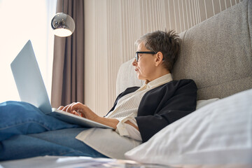 Adult businesswoman in glasses typing with laptop sitting on bed in hotel room