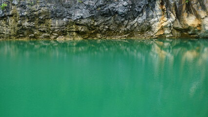 An image of a rocky cliff face reflected in the calm, emerald-green waters of a lake.