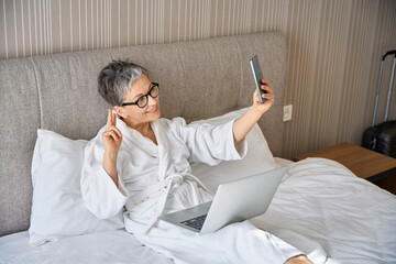 Happy adult woman making selfie on phone while sitting on bed with laptop in hotel