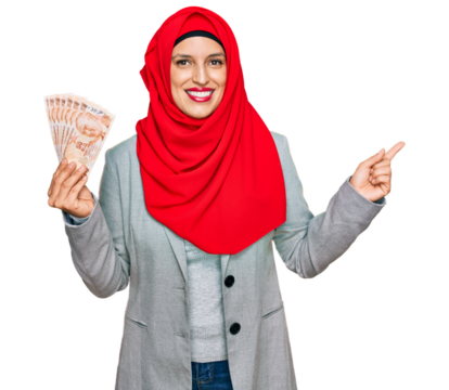 Beautiful hispanic woman wearing islamic hijab holding turkish lira banknotes smiling happy pointing with hand and finger to the side
