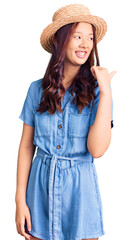 Young beautiful chinese girl wearing summer hat smiling with happy face looking and pointing to the side with thumb up.