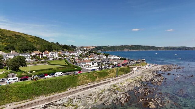 Drone footage over the Hannfore Beach showing coastal buildings, parked cars, road and green hills