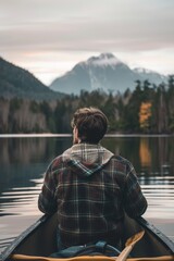 Fototapeta premium young man wearing a rugged flannel jacket and jeans sitting in a canoe on lake looking at distant mountains in background
