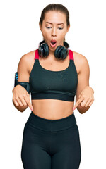 Beautiful blonde woman wearing gym clothes and using headphones pointing down with fingers showing advertisement, surprised face and open mouth