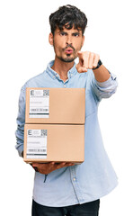 Young hispanic man holding delivery package pointing with finger to the camera and to you, confident gesture looking serious