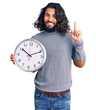 Young arab man holding big clock surprised with an idea or question pointing finger with happy face, number one